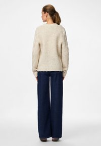 Χοντρό πλεκτό πουλόβερ με κρεμ χρώμα και υφή που δημιουργεί σχέδιο· άνετη εφαρμογή. Συνδυάζεται με ψηλόμεσα, σκούρα μπλε παντελόνια με φαρδιά μπατζάκια. Απλός στρογγυλός λαιμός.