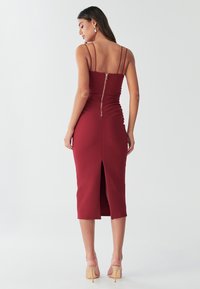 Žena ve formálním burgundském midi šatech bez rukávů s efektním zipem na zádech a rozparkem, doplněná béžovými sandály na vysokém podpatku, stojící na bílém pozadí.