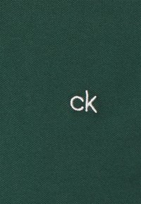 Grøn stof med en tekstureret overflade og et hvidt broderet "ck" logo. Designet er enkelt og fremhæver minimal branding.