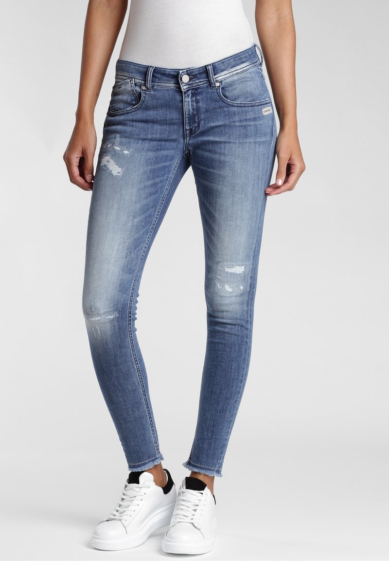 Vartalonmyötäiset, siniset kulutetut farkut repaleisilla reunuksilla, valkoiset tennarit ja istuva valkoinen paita mallilla.