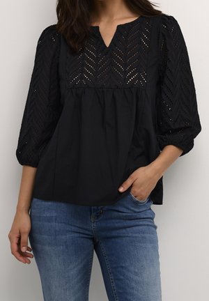 Vrouw draagt een zwarte blouse met chevron kantpatroon op mouwen en borst, gecombineerd met blauwe jeans, hand in de zak.