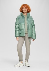Groene pufferjack met een glanzende afwerking, ribbelpanelen op de schouders, ritszakken en een hoge kraag. Draag het over een lichtgroene top en grijze jeans.