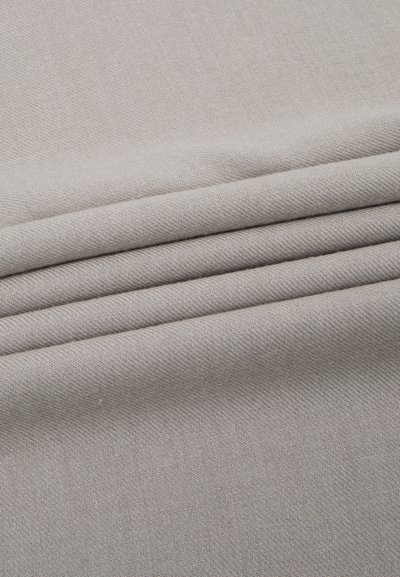 Tessuto di un grigio chiaro, caratterizzato da una superficie testurizzata con distintivi motivi diagonali e una leggera lucentezza, disposto in strati piegati.