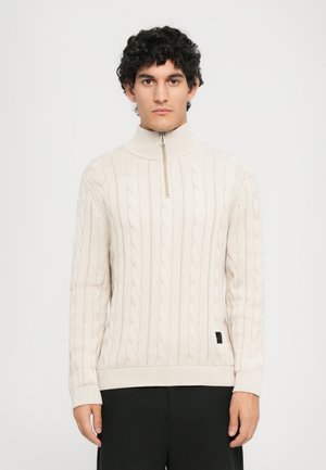 Hombre con cabello rizado vistiendo un suéter de punto en color crema con un cuello de cremallera corta y pantalones negros, de pie contra un fondo liso.