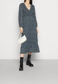 Robe midi à manches longues imprimée florale en noir et bleu, avec un décolleté en V et un ourlet volanté. Associée à un sac à main texturé blanc et des bottes noires.