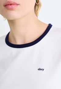 Balts T-krekls ar tumši zilu ribu apkakli un mazu izšūtu "obey" logo uz krūškurvja. Gluda kokvilnas auduma, cieši pieguļošs dizains.