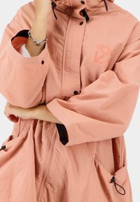 Růžová lehká bunda s vysokým límcem, velkými kapsami a zipem na přední straně. Má logo na hrudi a elastické stahovací šňůrky na kapuci.