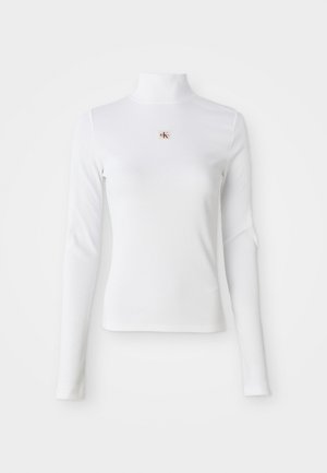 Garais piedurkņu baltais turtlenecks augšdaļa, izgatavota no ribota materiāla. Tam ir neliels izšūts logotips centrā. Pietuvojošs dizains bez rakstiem.