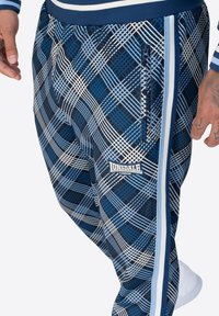 Mėlynos ir baltos languotos kelnės su užtrauktuku, elastinga juosta ir šoninėmis juostelėmis. Priekinėje dalyje matomas Lonsdale logotipas.