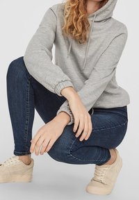 Sudadera gris con capucha ajustable y puños acanalados, combinada con jeans azul oscuro y zapatillas beige. Se puede ver la textura suave de la tela.