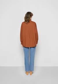 Camisa castanha de manga longa com um corte relaxado, bainha curvada e pregas detalhadas nas costas, combinada com jeans de perna larga em azul claro e sapatos castanhos claros.
