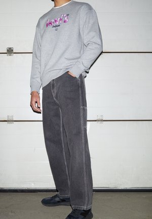 Person iført en grå Levi's sweatshirt med pink logo, mørkegrå jeans, sorte sneakers, står foran en hvid garagedør.