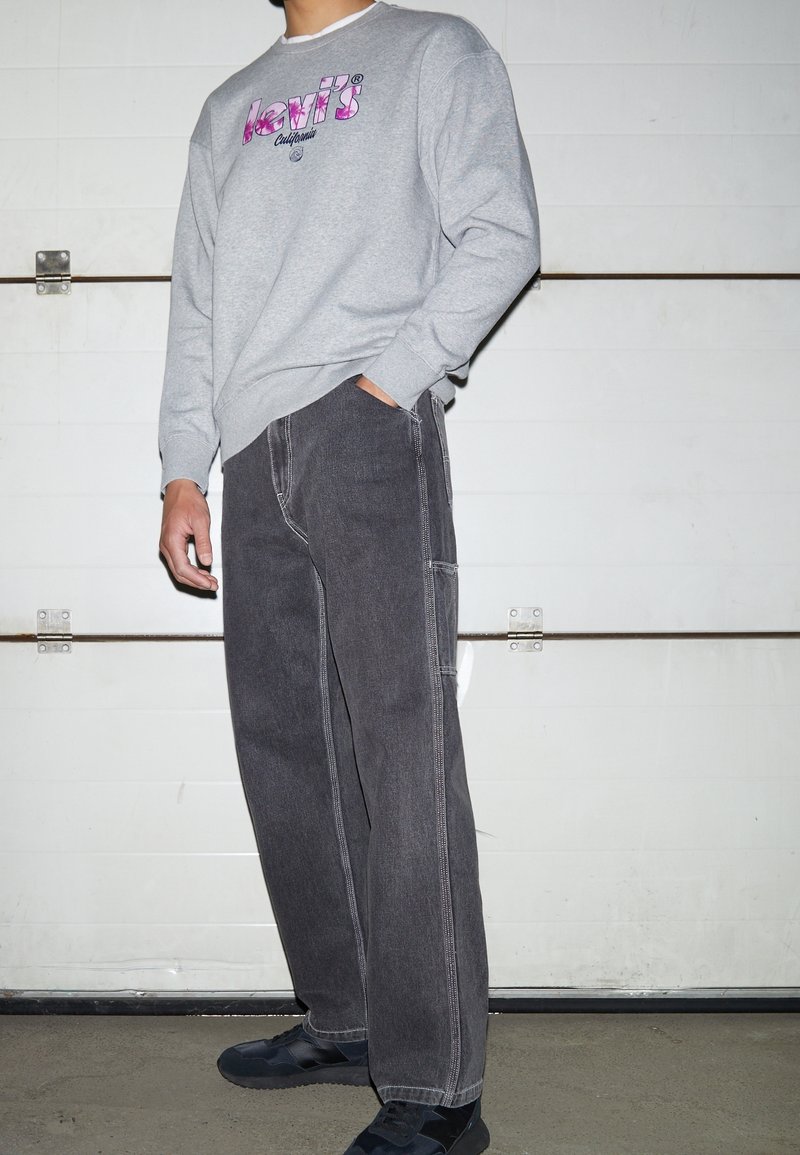 Personne portant un sweat-shirt gris Levi's avec logo rose, jean gris foncé, baskets noires, debout devant une porte de garage blanche.