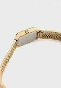 Ρολόι χειρός από χρυσό μέταλλο με ορθογώνια θήκη και πλεκτό βραχιόλι, εμφανιζόμενο από την πίσω πλευρά σε λευκό φόντο.