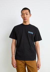 Svart bomull t-skjorte med korte ermer og rund hals, med en blå logo på venstre brystområde. Kombinert med brune bukser.