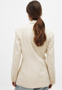 Blazer beige ajusté avec une texture lisse, des épaules structurées et des accents à trois boutons sur les manches. Silhouette ajustée.