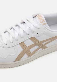 Weißer Sneaker mit beigen Akzenten, ausgestattet mit einem strukturierten Leder-Obermaterial, flachen weißen Schnürsenkeln und einer Gummisohle mit Perforationen zur Belüftung.