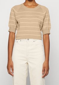 Person bærende en lys beige kortærmet sweater med tynde vandrette striber og højtaljede off-white fløjlbukser, der står imod en ensfarvet baggrund.