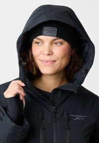 Black waterproof jacket with a hood, featuring zippered pockets, a logo patch, and thumbholes. Worn with a black knit beanie.