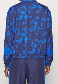 Mörkblå vindjacka med geometriska blå mönster, elastiska muddar och fåll, med hög krage och kontrasterande blå panelaccents.