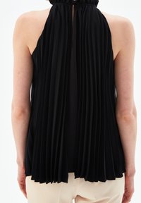 Ärmlös svart plisserad topp med öppning i ryggen och knytkrage, iförd med ljusbeiga byxor, visad bakifrån.