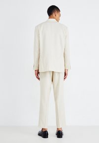 Traje de color beige claro que incluye una chaqueta ajustada con una única abertura y pantalones a juego, fabricado en tejido texturizado. Zapatos negros completan el conjunto.