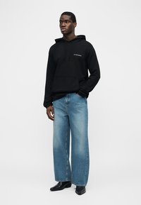 Junger Mann steht und trägt einen schwarzen Hoodie mit dem Schriftzug "LES BENJAMINS", hellblaue Jeans mit weitem Bein und schwarze, spitze Stiefel.