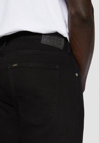 Gros plan d'une personne portant un jean noir Lee avec un patch logo et une chemise blanche, montrant la poche arrière et une partie du bras.
