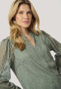 Blusa verde menta con un intricato pizzo a motivo diamantato, caratterizzata da un collo a chiave e maniche a palloncino trasparenti con bordo in pizzo.