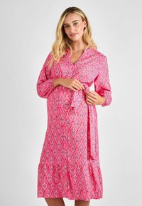 Robe de maternité rose à motif avec des manches longues, décolleté en V et taille nouée. Présente un design géométrique en blanc. Tissu léger.