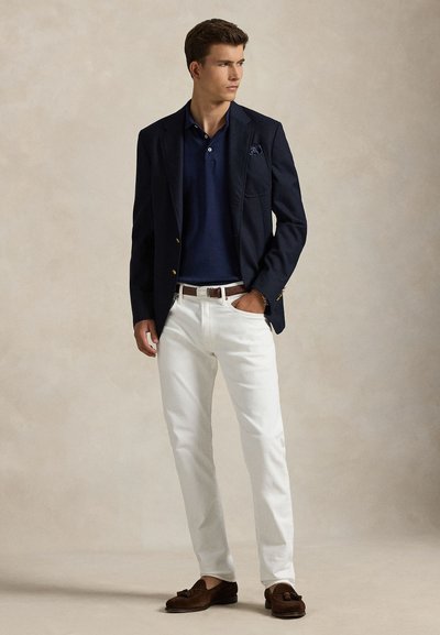 Junger Mann trägt einen marineblauen Blazer, ein marineblaues Polohemd, weiße Hose, braunen Gürtel und braune Tassel-Loafers, steht mit einer Hand in der Tasche.