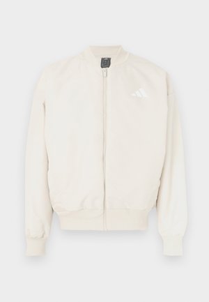 Beige bomberjakke med glidelås foran, ribbestrikkede mansjetter og kant, samt en liten hvit logo på venstre bryst. Glatt stofftekstur.