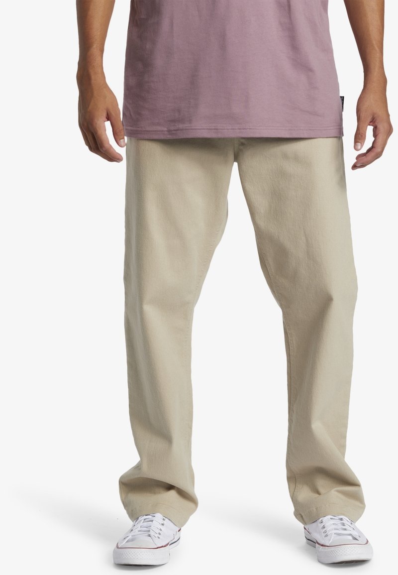 Homme portant un pantalon beige droit, des baskets blanches et un t-shirt mauve, debout, les bras détendus le long du corps, sur fond blanc.