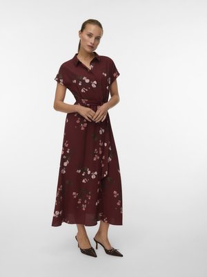 Woman standing wearing a burgundy midi dress with short sleeves, collar, floral pattern, waist tie, and dark pointed-toe heels.