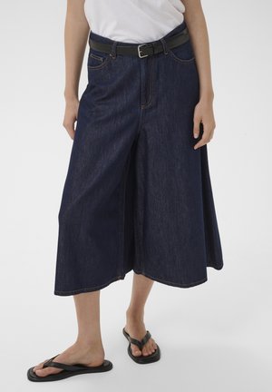 Naine kannab tumel siniseid laia säärega teksapükse, musta vööd, valget topi ja musti plätusid, seisab ühevärvilise valge tausta ees.