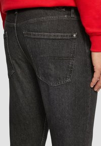Jean en denim noir avec une coupe slim, présentant des poches arrière doublées et des détails de couture subtils. Le tissu a une finition lisse et légèrement texturée.