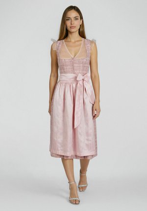 Frau trägt ein ärmelloses pinkes Dirndl mit tailliertem Mieder, transparenten Tupfen-Tüll-Ärmeln und einer Satin-Schürze, die in der Taille zu einer Schleife gebunden ist.