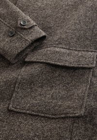 Brun hørnet vælg uldfrakke med jakkeslag, sorte knapper og en brystlomme. Skygget tekstur med en varm, blød finish.