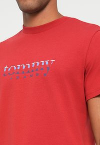 Camiseta de algodón roja con cuello redondo, que presenta un estampado gráfico azul de "tommy JEANS" en el pecho. Mangas cortas, textura suave.