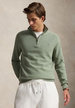 Mladi muškarac nosi svijetlozeleni džemper s četvrtinskim patentnim zatvaračem preko bijele košulje i bijelih hlača s vezicama, gleda dolje s rukama blizu džepova.