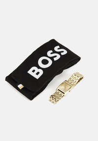 Auksinis metalinis laikrodis su stačiakampiu ciferblatu ir sujungta apyranke, šalia juodos audinio dėžutės, ant kurios yra balti užrašai "BOSS".