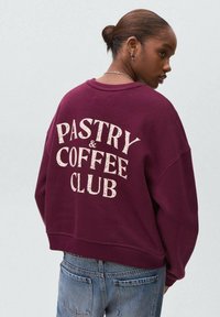 Giovane donna con capelli castani raccolti in un bun, che indossa una felpa bordeaux con la scritta "PASTRY & COFFEE CLUB" e jeans blu, rivolta di lato.