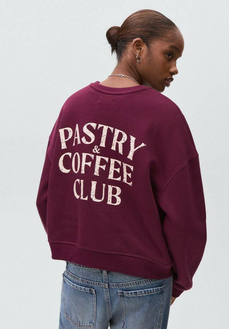 Giovane donna con capelli castani raccolti in un bun, che indossa una felpa bordeaux con la scritta "PASTRY & COFFEE CLUB" e jeans blu, rivolta di lato.