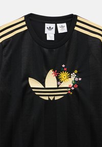 Μαύρο μπλουζάκι Adidas με χρυσό σχέδιο τριών ραβδώσεων στους ώμους και κεντήματα με λουλούδια γύρω από το εμβληματικό λογότυπο. Το ύφασμα έχει υφή με ανάγλυφο φινίρισμα.