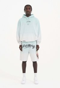 Lyseblå hættetrøje med en frontlomme og grafisk detalje. Matchende shorts har fed, sort skrift og farverigt print på siden. Hvide sneakers.