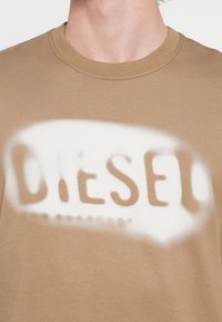 Καφέ βαμβακερό t-shirt με λευκό σπρέι που σχηματίζει το λογότυπο "DIESEL" στο στ�ήθος, με στρογγυλή λαιμόκοψη και διακριτή υφή.