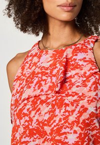 Blusa senza maniche a fantasia rossa e rosa con volant diagonale sul collo, realizzata in tessuto leggero. Accento con collana in metallo lucido.