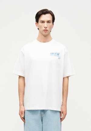 Muž v bielom oversized tričku s modrým nápisom "Couture Beach Club" na hrudi, v svetlo modrých rifliach, stojaci na jednoduchom pozadí.