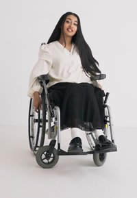 Una persona in sedia a rotelle indossa una blusa crema chiaro e una gonna nera plissettata. La sedia a rotelle ha grandi ruote posteriori e un poggiapiedi. L'ambientazione è neutra.