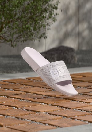 Biały teksturowany sandał na płaskiej podeszwie z logo "BOSS" unoszącym się nad drewnianą deską i kamienistą powierzchnią na zewnątrz.